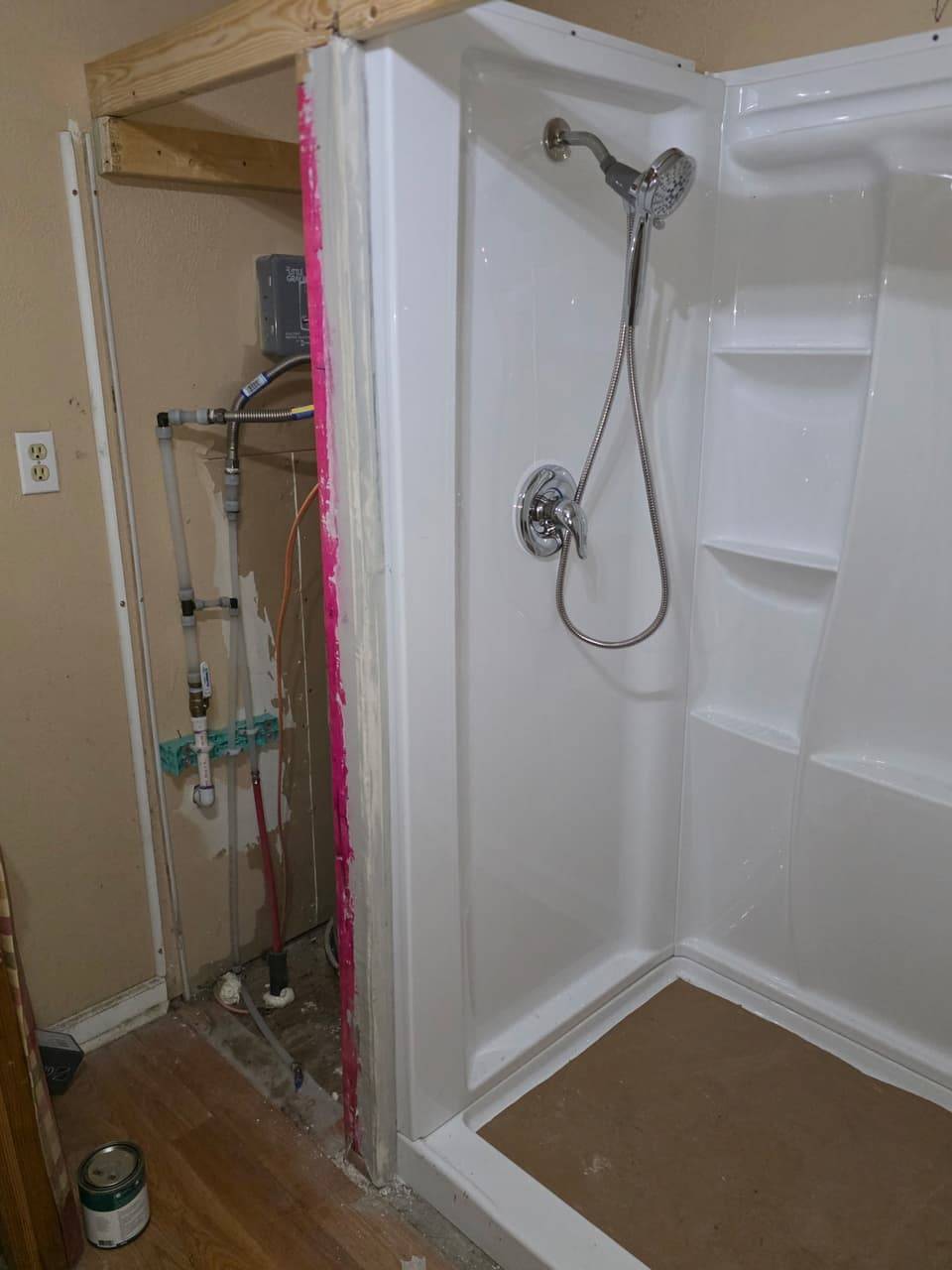 New white shower surround with a handheld shower head beside a framed utility closet with supply lines and a shutoff valve